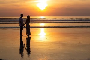 The silhouette of a couple and their reflection in the sand on a beach at sunset.