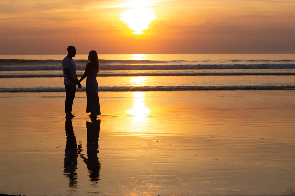 The silhouette of a couple and their reflection in the sand on a beach at sunset.