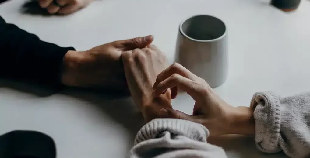 Close-up of a man and a woman holding hands across a white table with coffee mugs, symbolizing emotional support and vulnerability in a relationship.